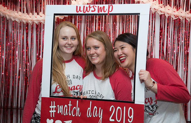 A group of three women stand in front of a frame for a photo that says #match day 2019