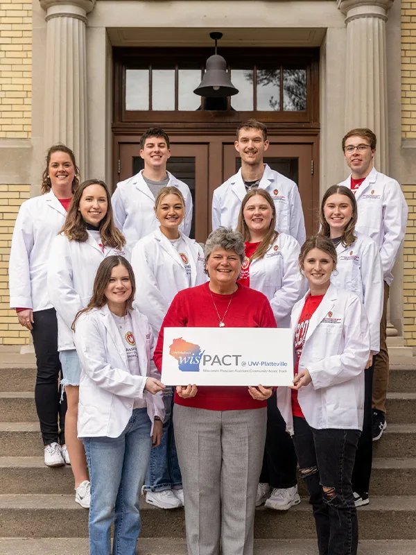 A group of students wearing white coats happily posing for a picture with a woman holding a "wisPACT@UW-Platteville Program" sign