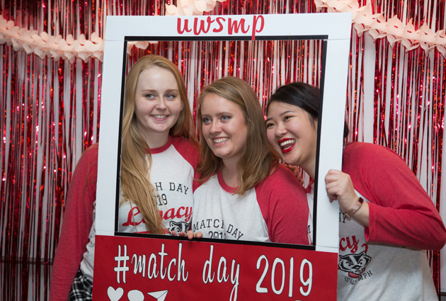 A group of three women stand in front of a frame for a photo that says #match day 2019