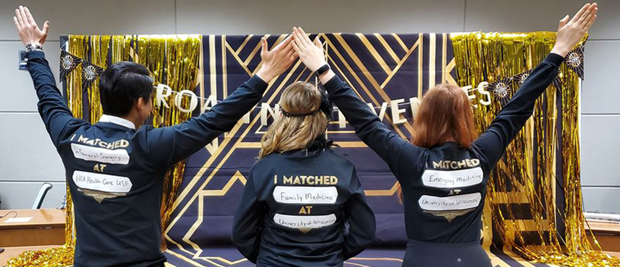 Three individuals stand with their backs to the camera and their arms in the air to celebrate a virtual Match Day event in 2020