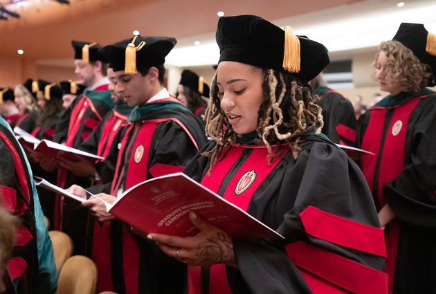 A woman wearing a cap and gown looks at a printed publication during a graduation event