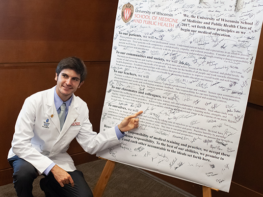 A man in a white coat points to a signature on a large poster