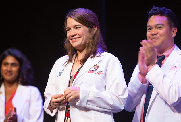 A woman smiles while on a stage wearing a white medical coat