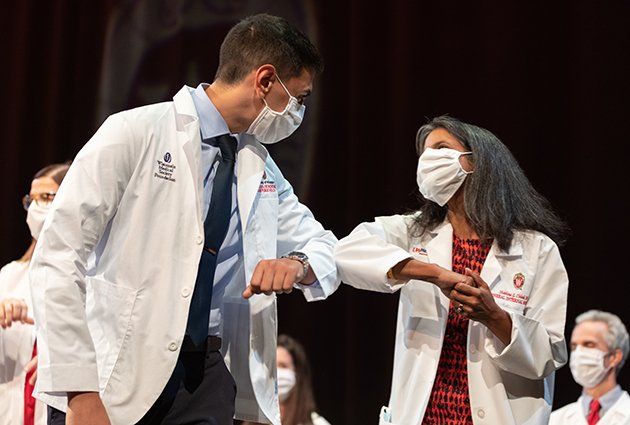 Two people wearing white medical coats tap elbows while wearing masks during a ceremony