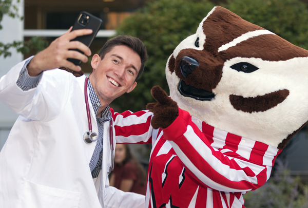A med student, wearing a white coat smiles and takes a photo with Bucky Badger