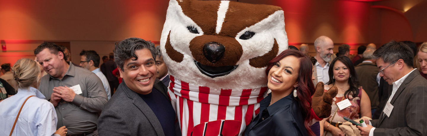 Two people smiling with Bucky Badger at a reunion event