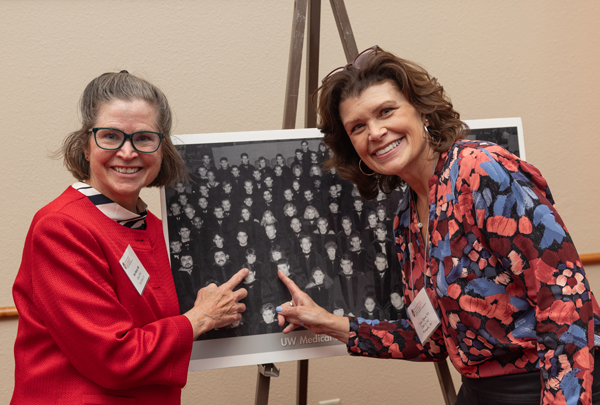 Two women smile next to a class photo