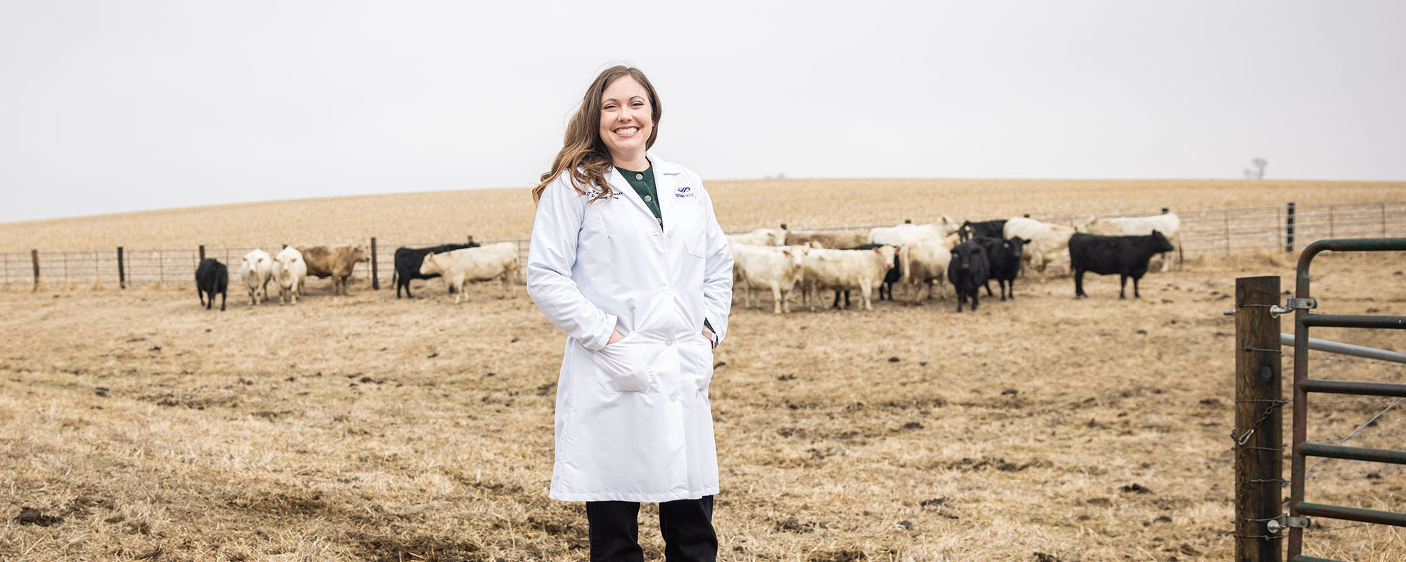Kelsey Schmidt wearing a doctor's white coat on a farm with cows grazing behind her.