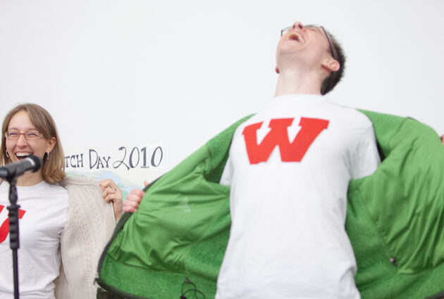 A man and a woman stand together, the woman has a red letter u on her white shirt and the man has q red letter w on his white shirt.