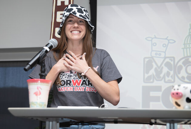 A woman stands at a podium wearing a cow print hat at a Match Day event in 2024
