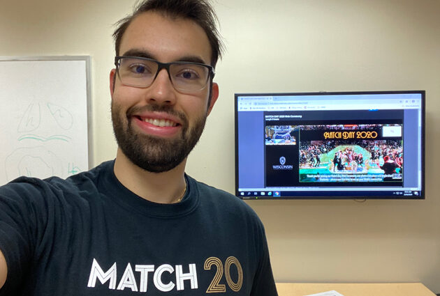 A man smiles while wearing a Match Day 2020 black t shirt and standing in front of a television screen
