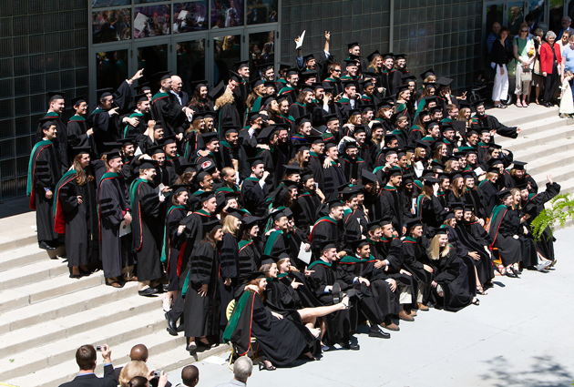 A large group of graduates sit on the stairs wearing their caps and gowns