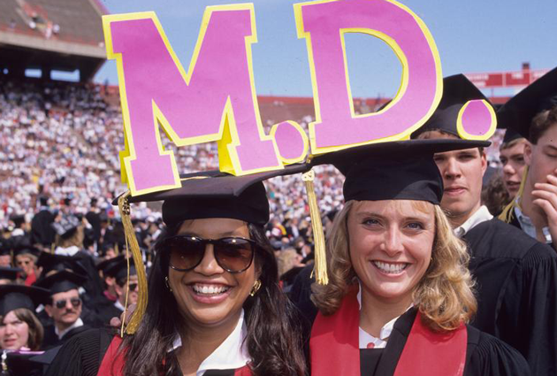 Two women wearing caps and gowns at a graduation ceremony with the letters "M" and "D" above their heads