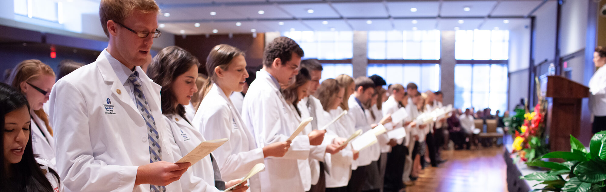 A large group of people wearing white medical coats read from papers