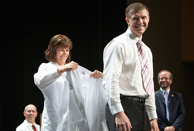 A woman puts a white medical coat on a man during the white coat ceremony
