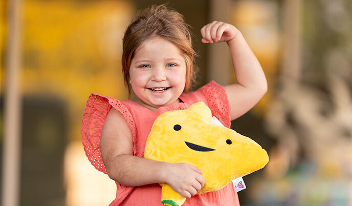 Transplant recipient Lucy Sughroue smiles while flexing her muscles and holding a stuffed toy