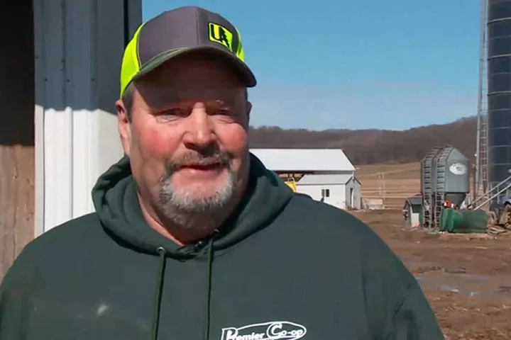 A man in a hoodie and baseball cap with a rural area behind him