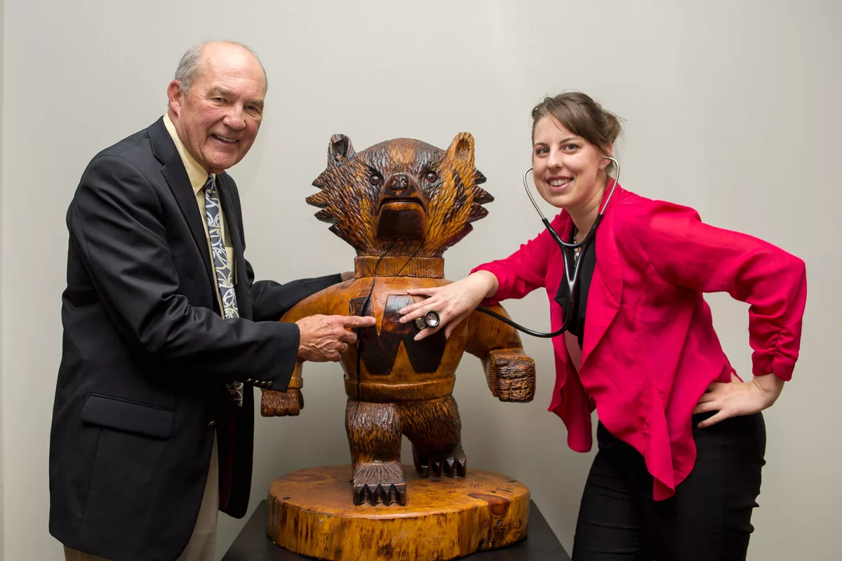 An alumni and a medical student posing with a wood carving of Bucky Badger, the medical student holding their stethoscope up to Bucky's heart